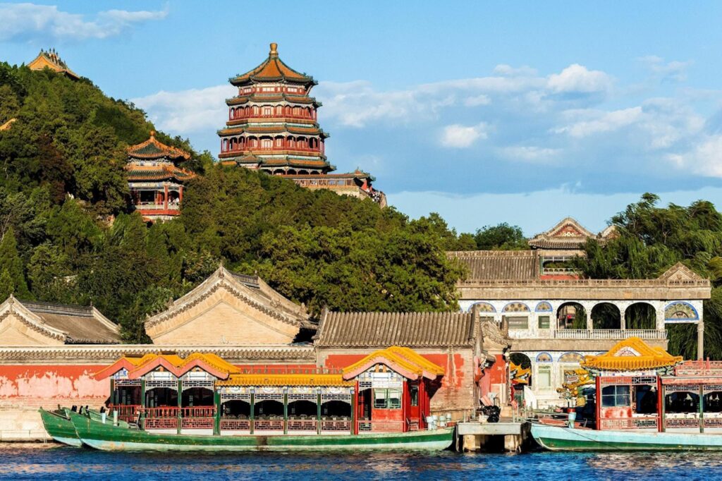 This image captures the grand Tower of Buddhist Incense perched atop Longevity Hill at the Summer Palace in Beijing. Traditional dragon boats and ornate pavilions line the edge of Kunming Lake in the foreground under a bright, clear sky.
