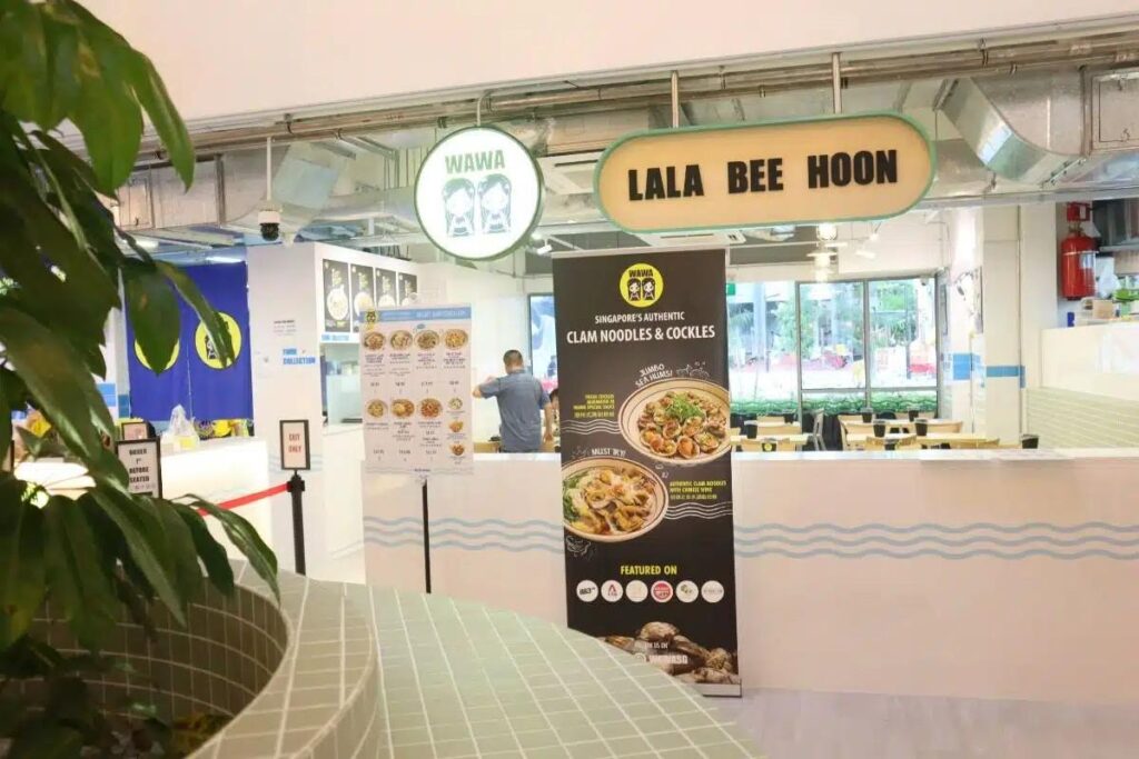 The image displays the bright, clean storefront of Wawa Lala Bee Hoon, featuring a white counter decorated with blue wave patterns and a tall banner advertising authentic clam noodles. Hanging above the entrance are circular and oval signs displaying the restaurant's name and mascot, while the interior reveals a minimalist dining area with light wood tables.