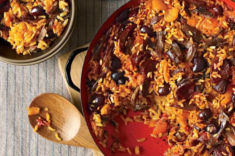 This top-down shot showcases a vibrant dish of rice mixed with tender pieces of duck and sweet apricots, served in a large red pan. A wooden spoon rests nearby on a striped tablecloth, while a smaller portion of the colorful meal is plated in a separate bowl.