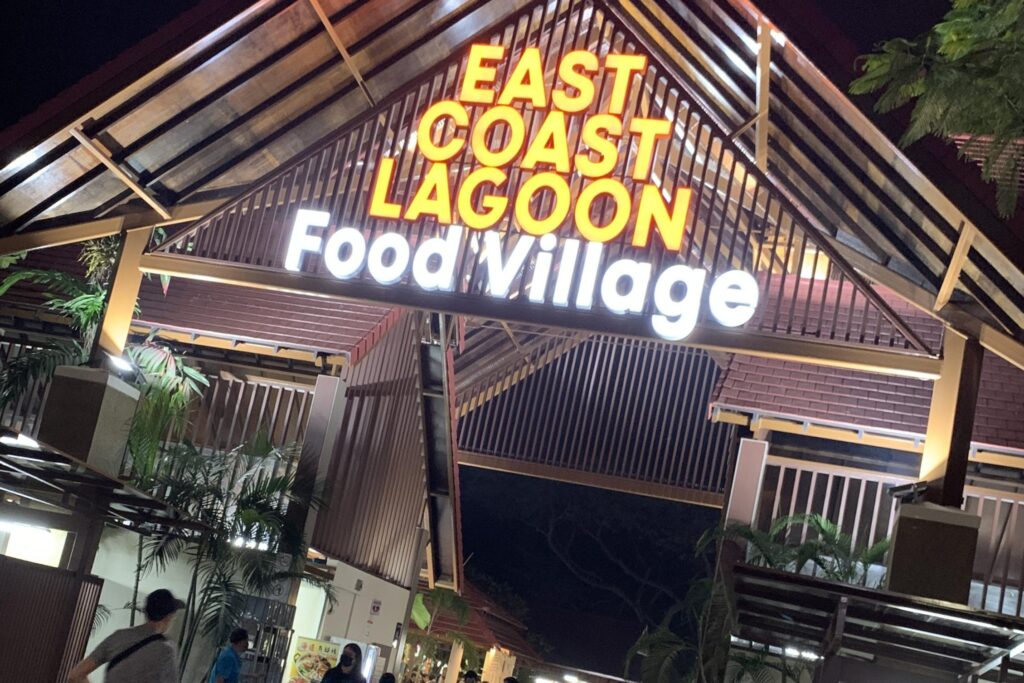 A brightly illuminated sign for the East Coast Lagoon Food Village hangs beneath the high, slatted wooden ceiling of an open-air pavilion at night. Tropical greenery and several visitors are visible near the entrance, which is framed by the traditional architectural style of the food centre.