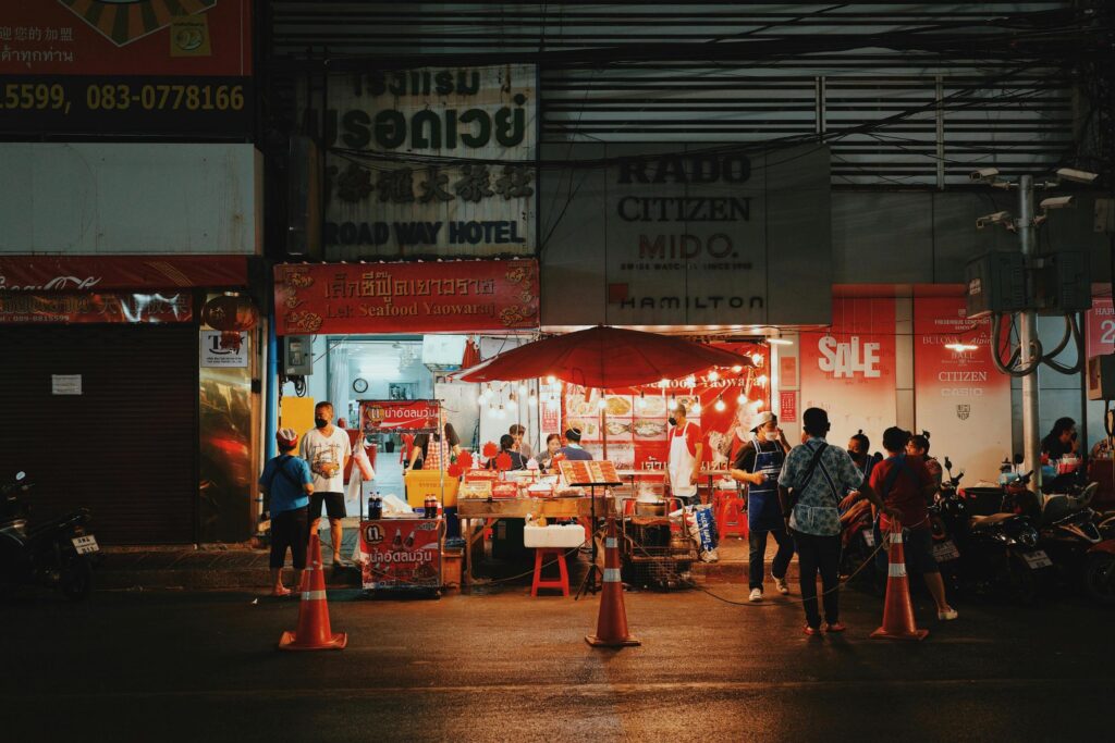 A brightly lit street food stall named "Lek Seafood Yaowaraj" operates at night under a large red umbrella, nestled between storefronts for a hotel and a watch shop. Customers and staff wearing masks congregate around the food preparation area, which is cordially blocked off from the street by orange traffic cones.