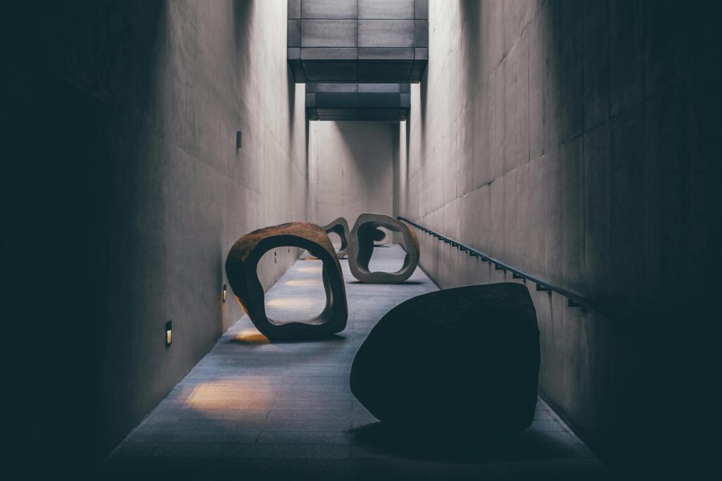 This narrow, modern corridor features high concrete walls and geometric overhead structures that let in slivers of natural light. Large, organic stone sculptures with hollowed-out centers are arranged along the path, creating a striking contrast between the architectural lines and natural forms.