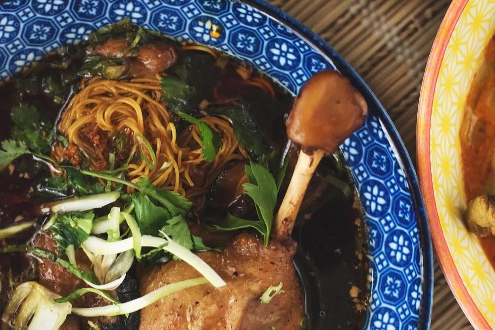 A roasted duck leg is served in a blue patterned bowl filled with a dark, savory broth and thin egg noodles. The dish is garnished with fresh herbs and sliced green onions, creating a comforting and flavorful presentation.