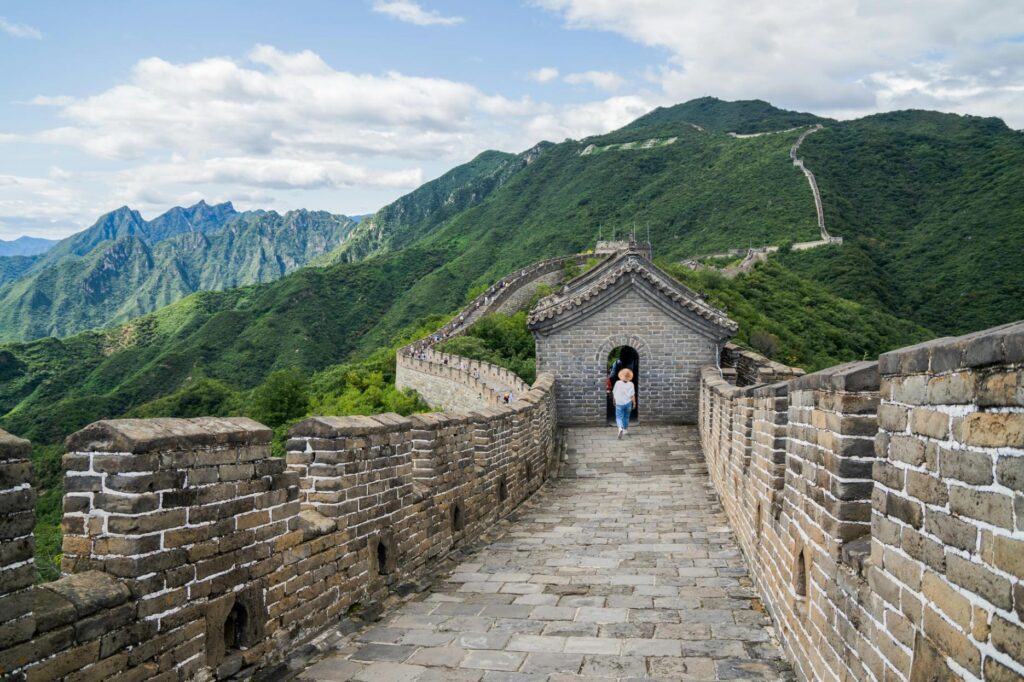 The Great Wall of China stretches across lush, green mountain ridges under a bright, cloudy sky. A stone watchtower stands in the foreground, with a lone traveler walking through its arched entrance along the ancient paved path.