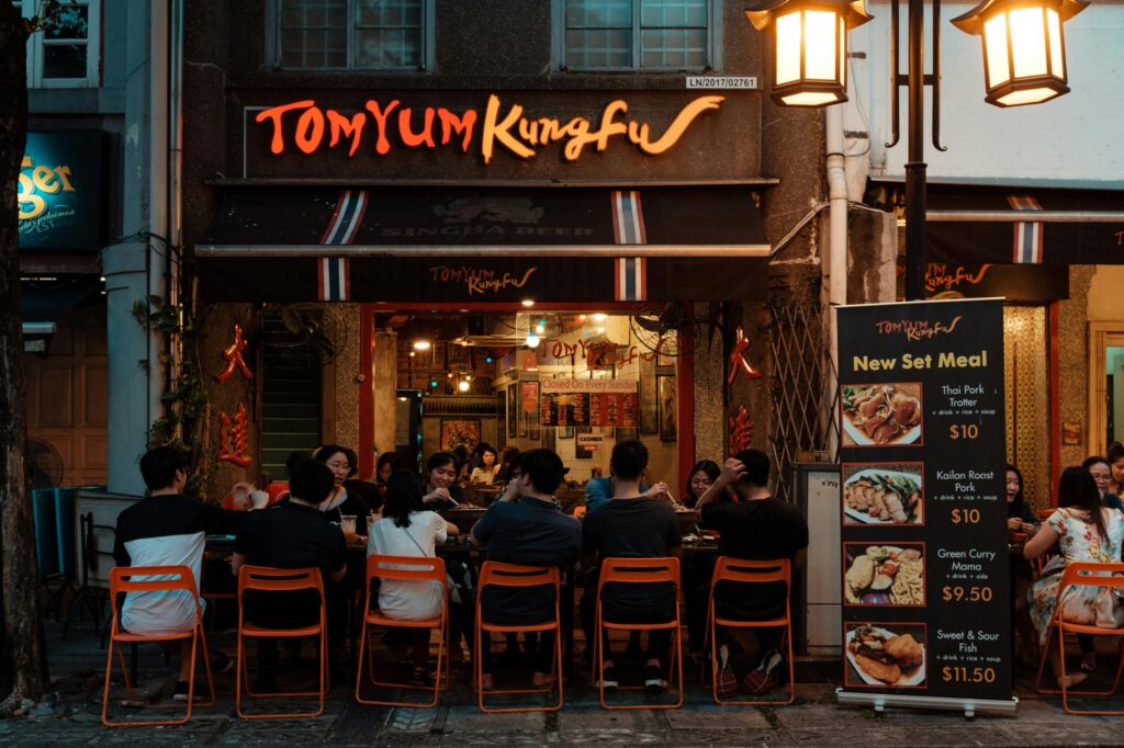 Patrons sit at outdoor tables under the glowing "Tom Yum Kungfu" sign of a lively Thai restaurant at night. A large menu board stands to the right, showcasing set meal specials like Thai Pork Trotter and Green Curry Mama for passersby.
