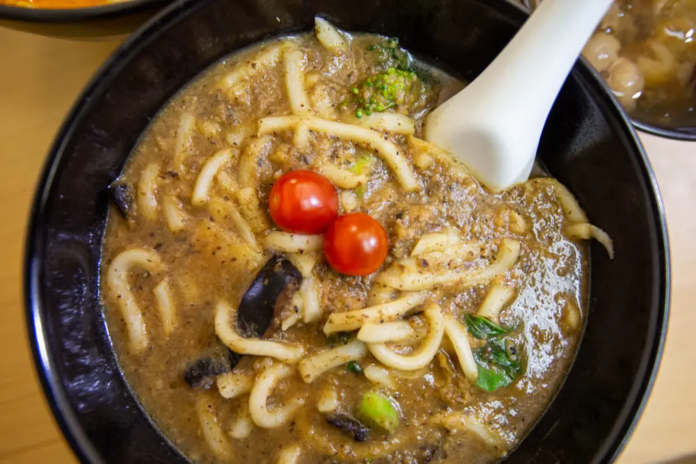 A black bowl is filled with thick udon noodles submerged in a savory, dark mushroom gravy topped with two bright cherry tomatoes. A white ceramic spoon rests in the dish, which is garnished with bits of broccoli and greens.