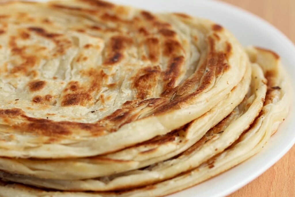 This close-up shot features a stack of golden-brown roti prata layered on a white plate, highlighting the bread's characteristic flaky and crispy texture. The circular flatbreads show distinct swirled patterns and toasted spots, suggesting they have been freshly cooked on a griddle.