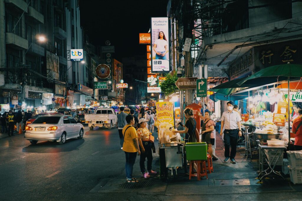 A bustling city street at night is filled with moving cars, glowing neon signs, and various street food vendors lining the sidewalks. Pedestrians navigate the busy area, while a brightly lit tall billboard and numerous shop banners add to the vibrant, urban atmosphere.