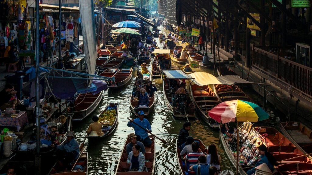 A bustling canal is packed with traditional wooden long-tail boats carrying tourists and fresh produce through a vibrant floating market. Vendors under colorful umbrellas line the narrow waterway, which is shaded by the rustic wooden structures of the surrounding docks.