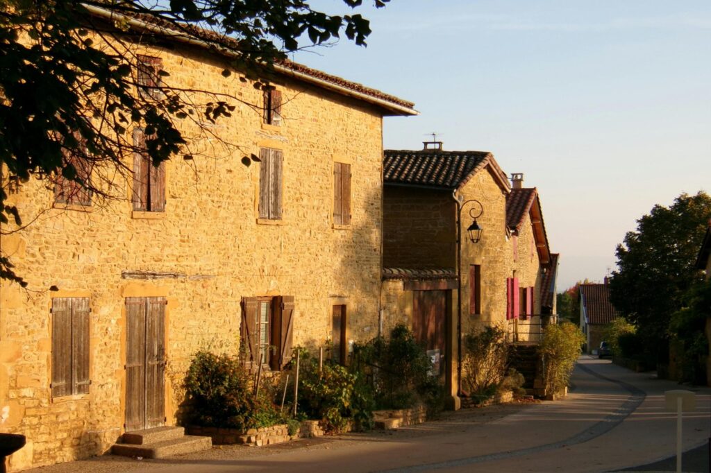 This narrow street is lined with historic golden-stone buildings featuring traditional wooden shutters and rustic tiled roofs. The warm, late-afternoon sun casts long shadows across the road as it curves gently toward a distant, hazy horizon.