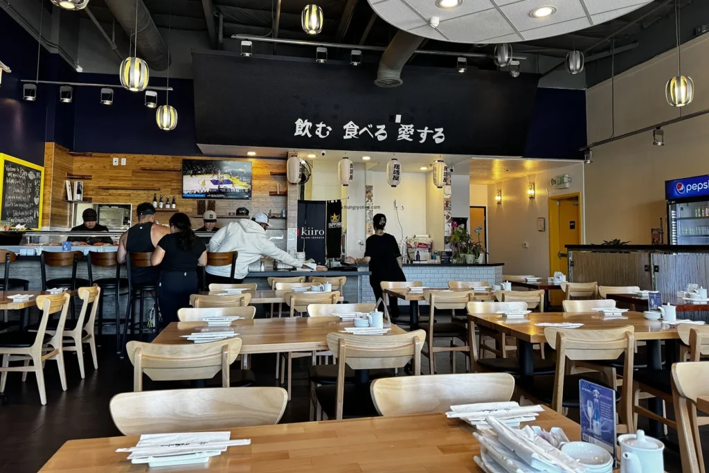 A modern Japanese restaurant interior features light wood tables and chairs arranged in a spacious dining area beneath a dark ceiling with stylish pendant lights. Several patrons are seated at a sushi bar in the background, which is anchored by a large black banner with white Japanese calligraphy.