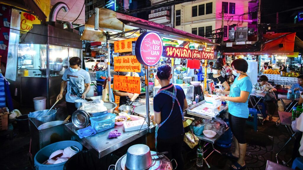 A brightly lit street food stall, featuring a red pig logo for "Nay Lek Uan Yaowarat," serves customers along a bustling sidewalk at night. Vendors wearing masks prepare dishes in large metal pots while patrons wait and dine at simple tables under the glow of neon signs.