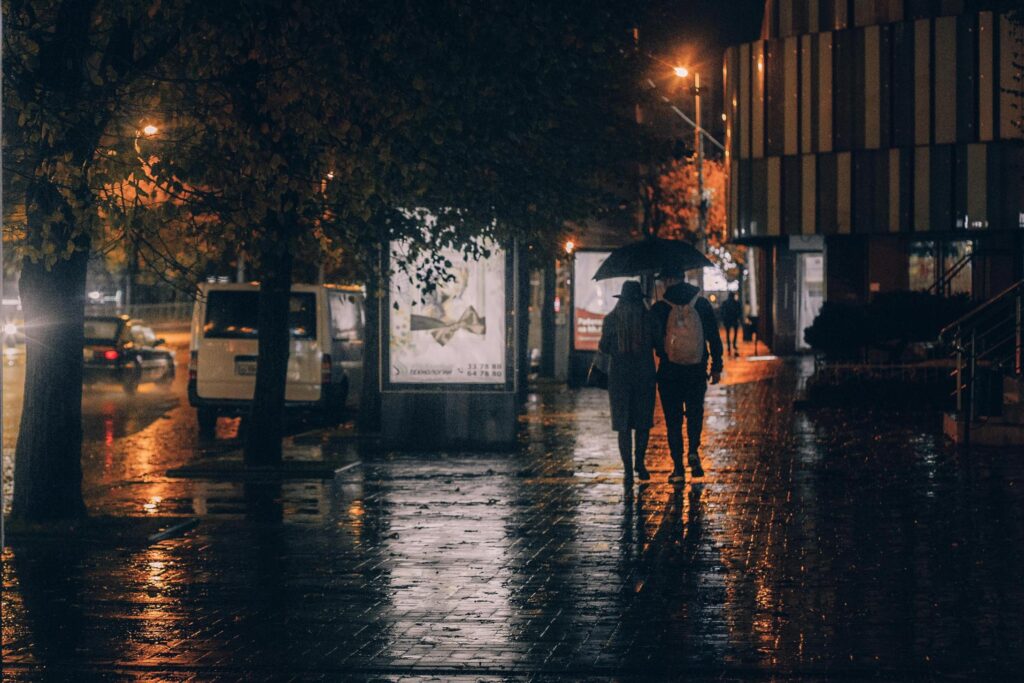 A couple walks together under a single umbrella along a rain-slicked city sidewalk at night. Warm streetlights and glowing advertisements reflect off the wet pavement, creating a moody atmosphere as they pass by parked vehicles and dark trees