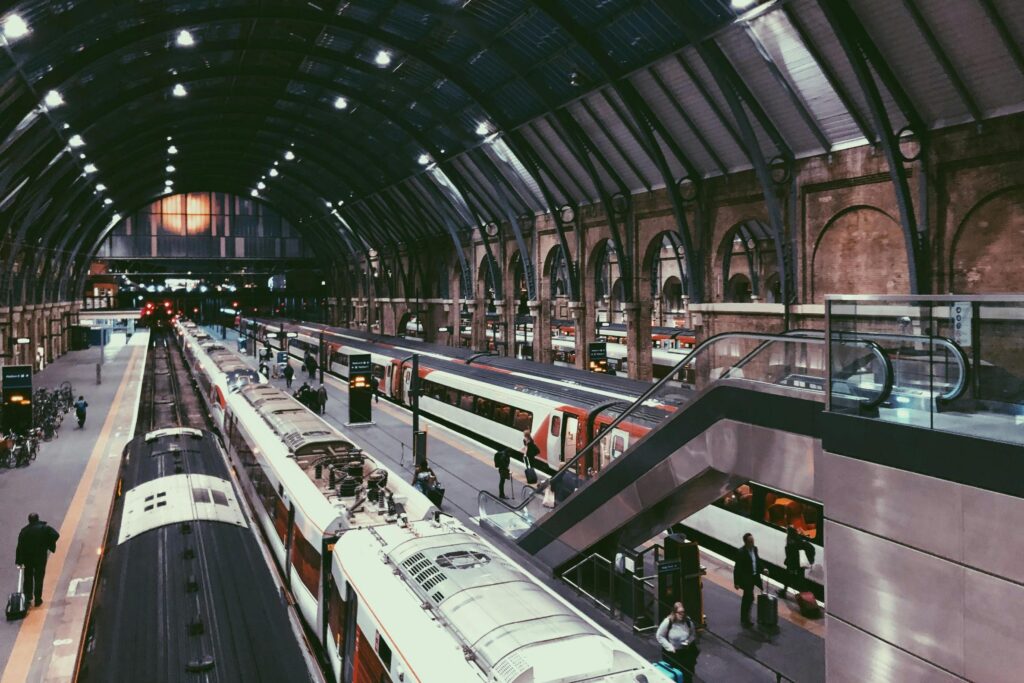 This image captures the bustling interior of a large, historic train station featuring a high vaulted glass ceiling and several long trains parked at platforms. Commuters with luggage are scattered throughout the station, navigating the space alongside a modern escalator and arched brick architecture