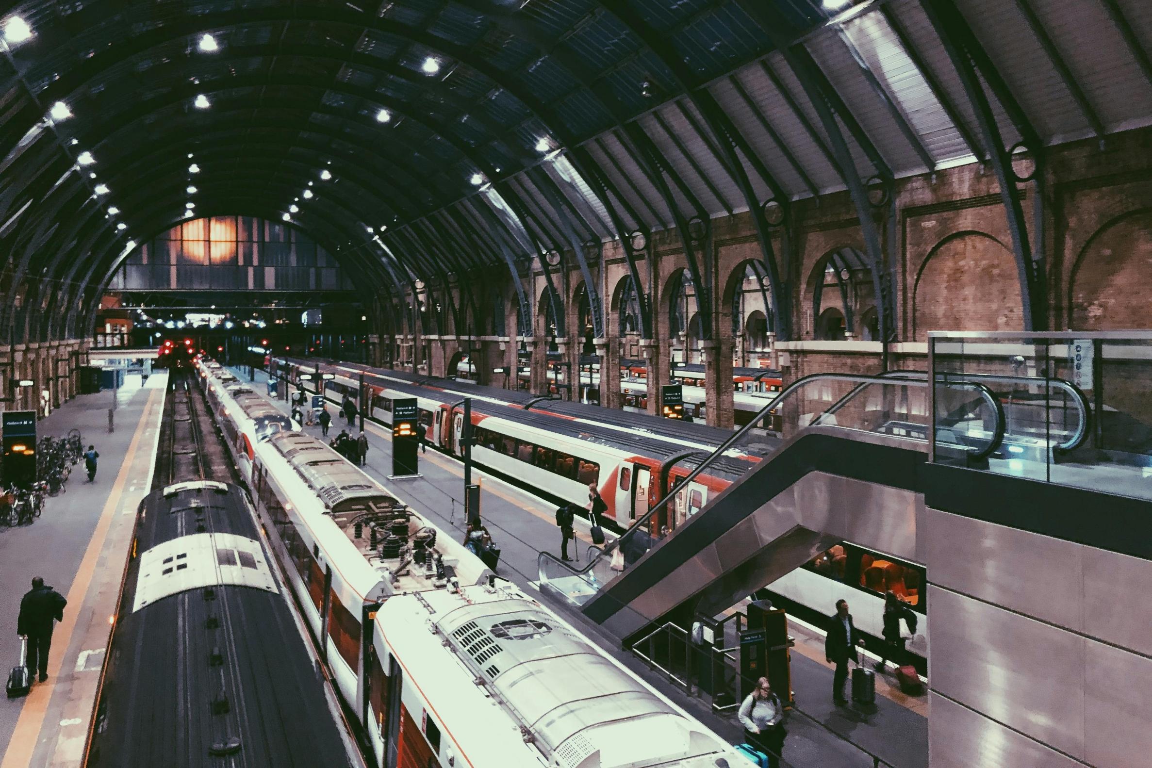 This image captures the bustling interior of a large, historic train station featuring a high vaulted glass ceiling and several long trains parked at platforms. Commuters with luggage are scattered throughout the station, navigating the space alongside a modern escalator and arched brick architecture