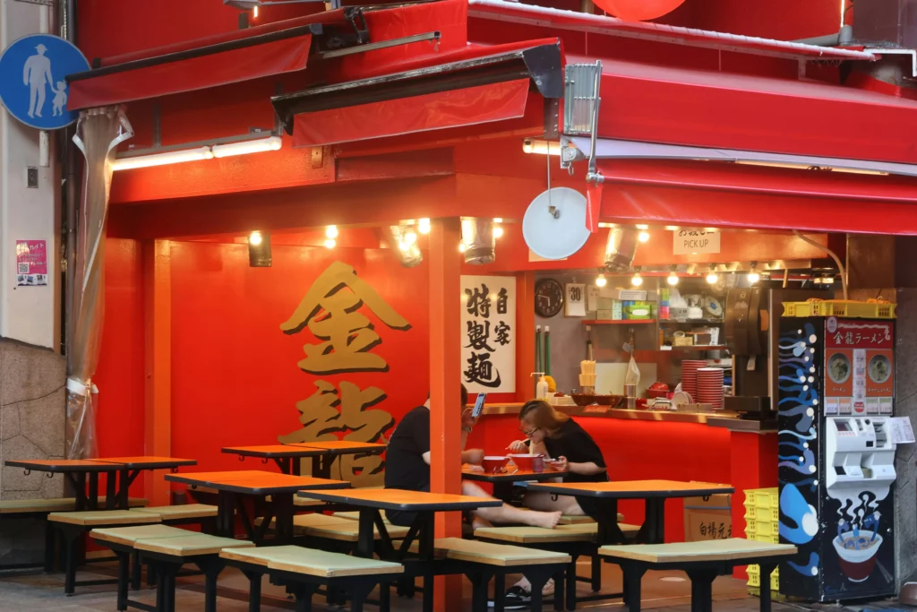 Customers eat at a vibrant, red-painted outdoor ramen shop featuring traditional Japanese signage and warm overhead lighting. The scene includes several empty wooden tables with green benches and a colorful vending machine for ordering on the side.