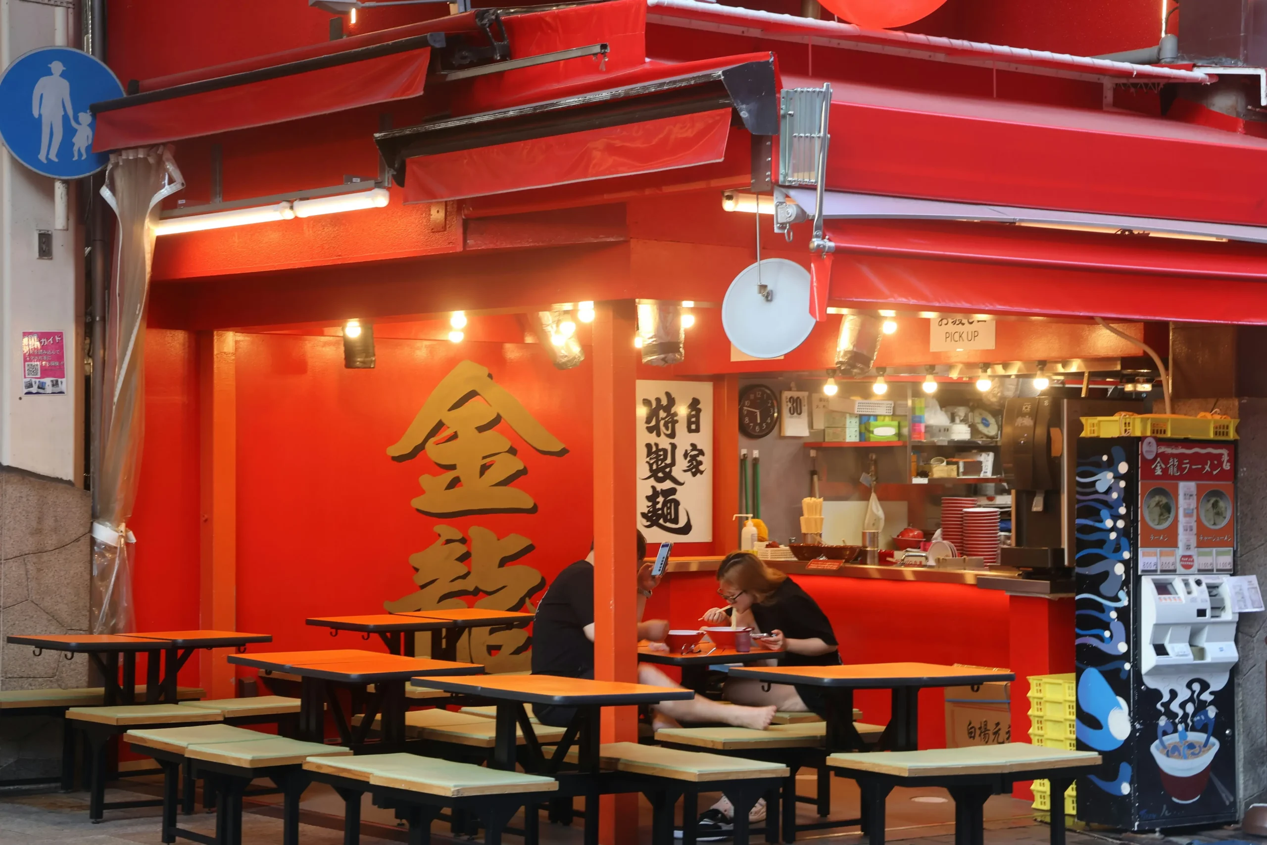 Customers eat at a vibrant, red-painted outdoor ramen shop featuring traditional Japanese signage and warm overhead lighting. The scene includes several empty wooden tables with green benches and a colorful vending machine for ordering on the side.
