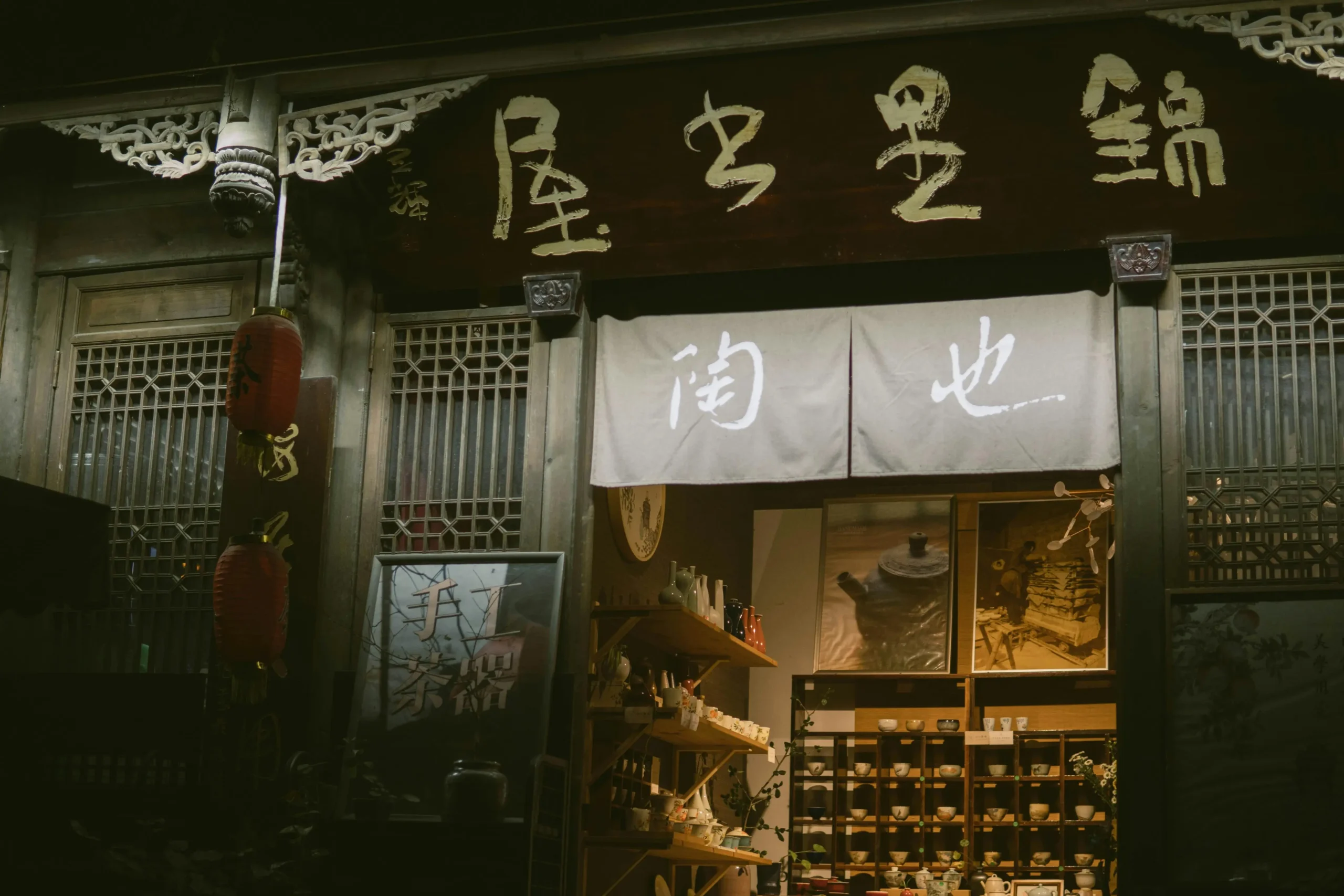 A rustic storefront in China features traditional wooden architecture and a large calligraphic sign hanging above the entrance. Through the open door, shelves of delicate ceramic teacups and pottery are illuminated by a warm, golden glow.