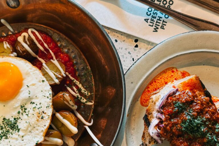 The image features a close-up overhead view of two savory brunch dishes, including a fried egg with roasted potatoes in a copper pan and a thick slice of topped toast on a ceramic plate. Both plates are garnished with fresh herbs and colorful sauces, set against a light-colored speckled tabletop with a white napkin and cutlery nearby.
