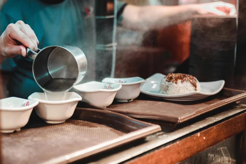 A cook pours steaming hot liquid from a metal measuring cup into several small white bowls lined up on a tray. In the background, a plate of white rice topped with a dark sauce sits ready for service in a busy kitchen environment.