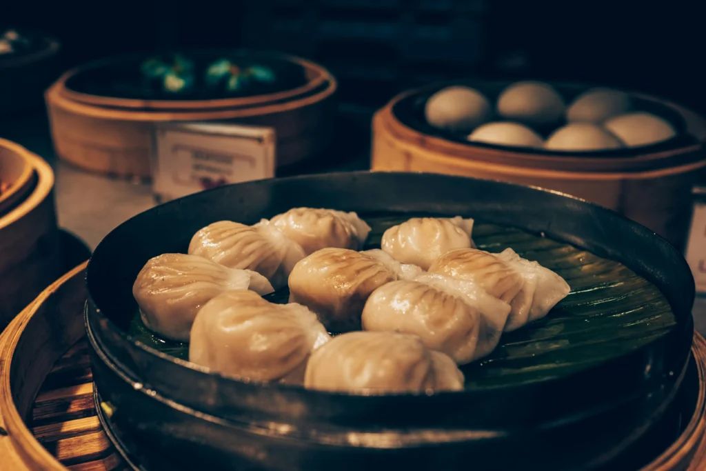 A variety of steamed dim sum, including translucent shrimp dumplings, are presented in traditional bamboo steamers lined with banana leaves. The warm, moody lighting highlights the textures of the delicate pleated wrappers and soft buns in the background.