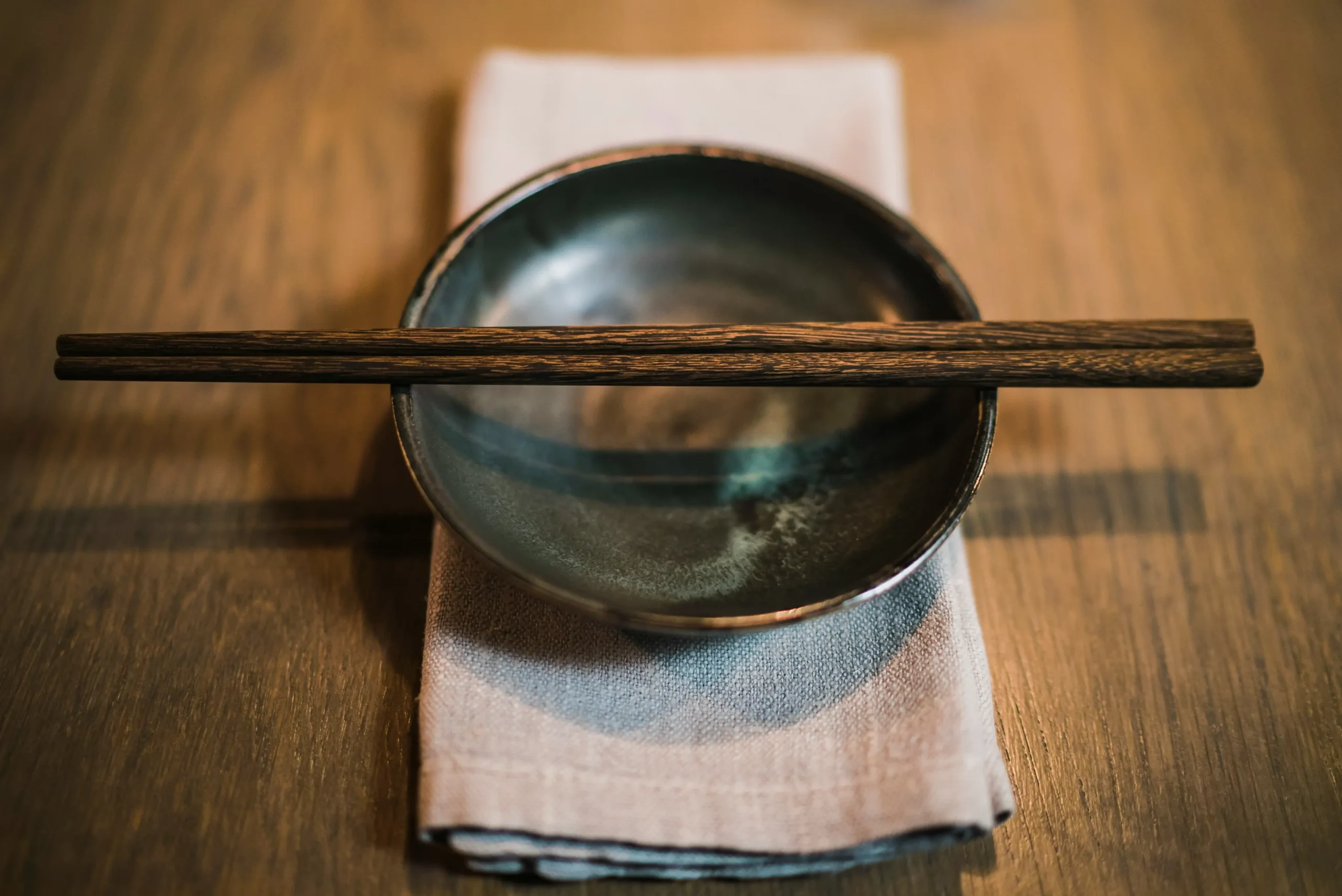 A pair of dark wooden chopsticks rests horizontally across the rim of a small, rustic ceramic bowl. The bowl is set atop a neatly folded light-colored napkin on a textured wooden table.