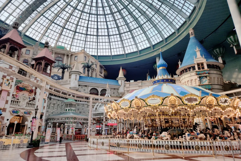 This indoor amusement park features a classic, illuminated carousel centered under a massive glass dome ceiling. Surrounding the ride are European-style buildings, castle turrets, and an elevated track that winds through the whimsical complex.