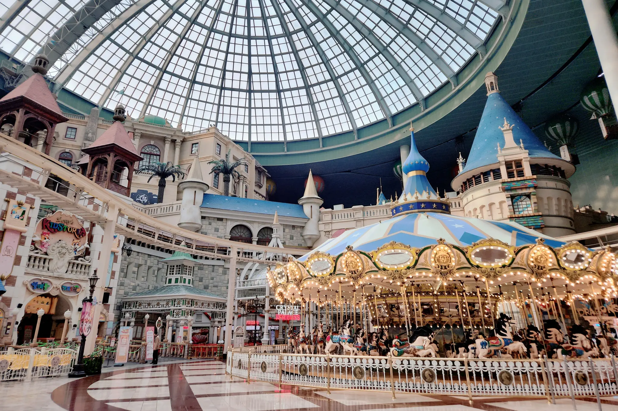 This indoor amusement park features a classic, illuminated carousel centered under a massive glass dome ceiling. Surrounding the ride are European-style buildings, castle turrets, and an elevated track that winds through the whimsical complex.