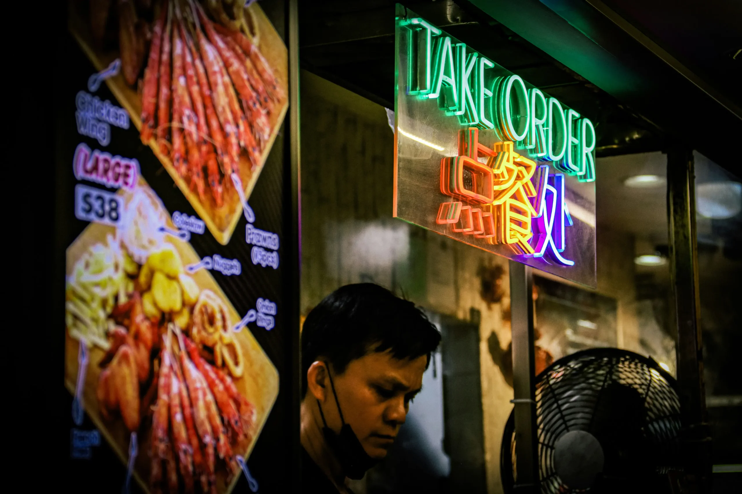 A man wearing a black mask stands behind a food stall counter, illuminated by a vibrant "TAKE ORDER" neon sign in English and Chinese. Beside him, a colorful menu board displays photos of fried seafood and snacks with prices listed.