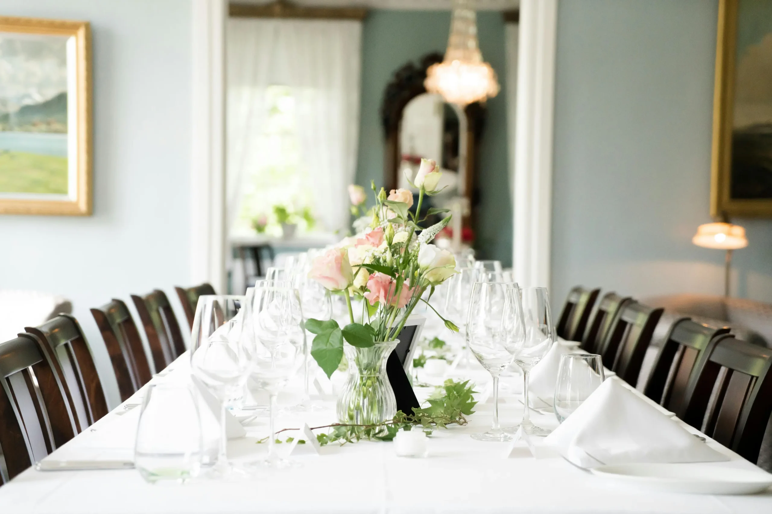 A long banquet table is elegantly set with white linens, crystal glassware, and delicate pink and white floral centerpieces in a bright, airy room. Dark wooden chairs line the table, leading the eye toward a glowing chandelier and a soft, blurred background of classical decor.