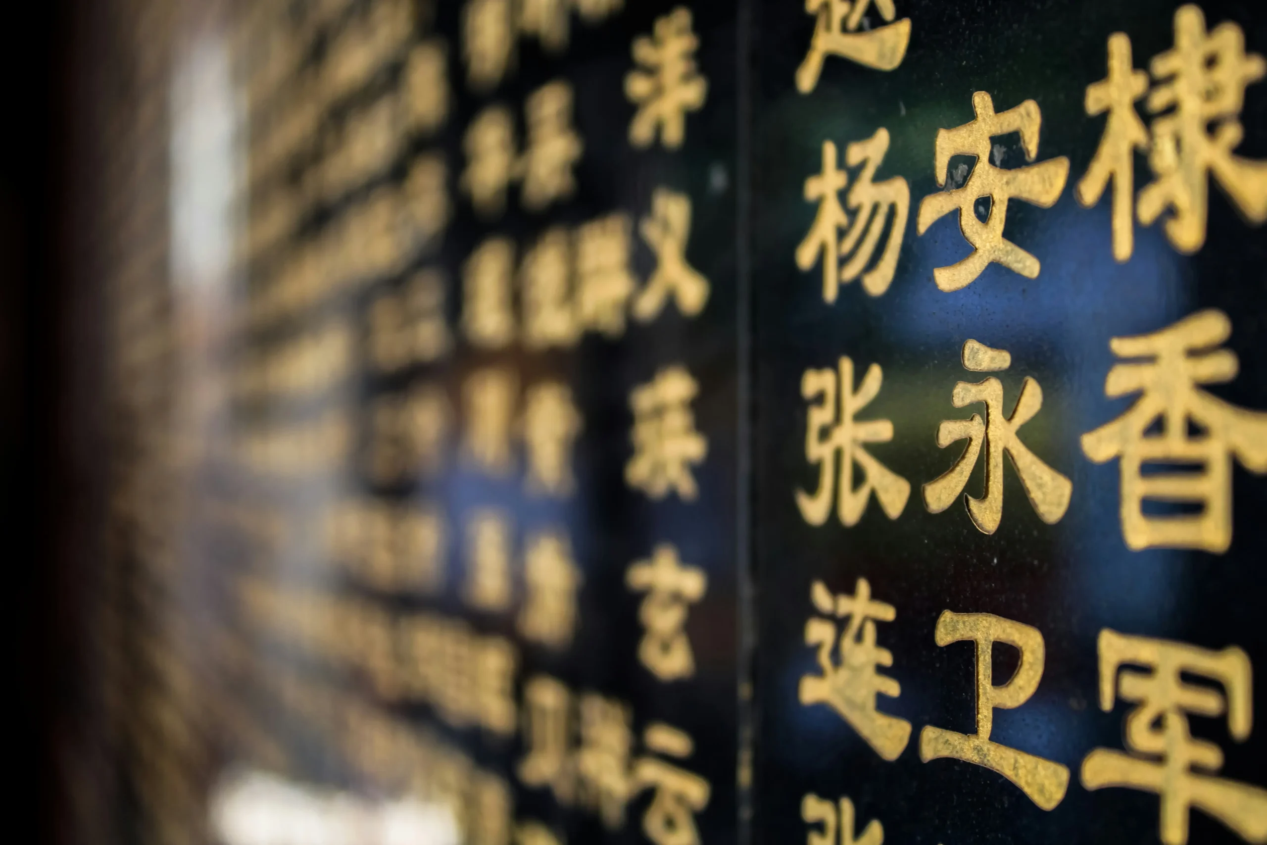 This image captures a close-up, shallow-depth-of-field shot of golden Chinese characters engraved onto a polished black stone surface. The foreground characters are sharp and legible, while the rest of the inscriptions blur into a soft, glowing bokeh effect as they recede into the distance.