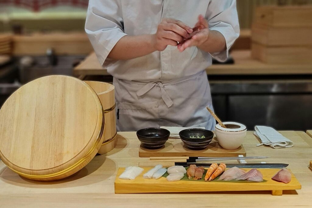 A sushi chef works behind a wooden counter, hand-pressing a piece of nigiri while a colorful assortment of fresh seafood sits prepared on a serving board in the foreground. The workspace is organized with traditional tools, including a large wooden rice tub, small condiment bowls, and long metal plating chopsticks.