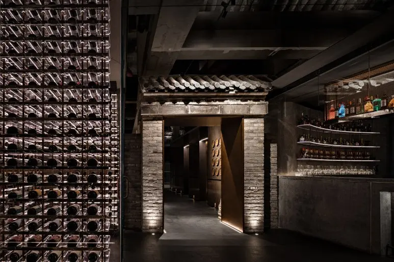 This upscale interior combines modern elements like an illuminated, floor-to-ceiling glass wine cellar and sleek bar with a traditional brick entryway topped by a tiled roof. The moody, low-light atmosphere creates a sophisticated contrast between contemporary design and classic architectural features.
