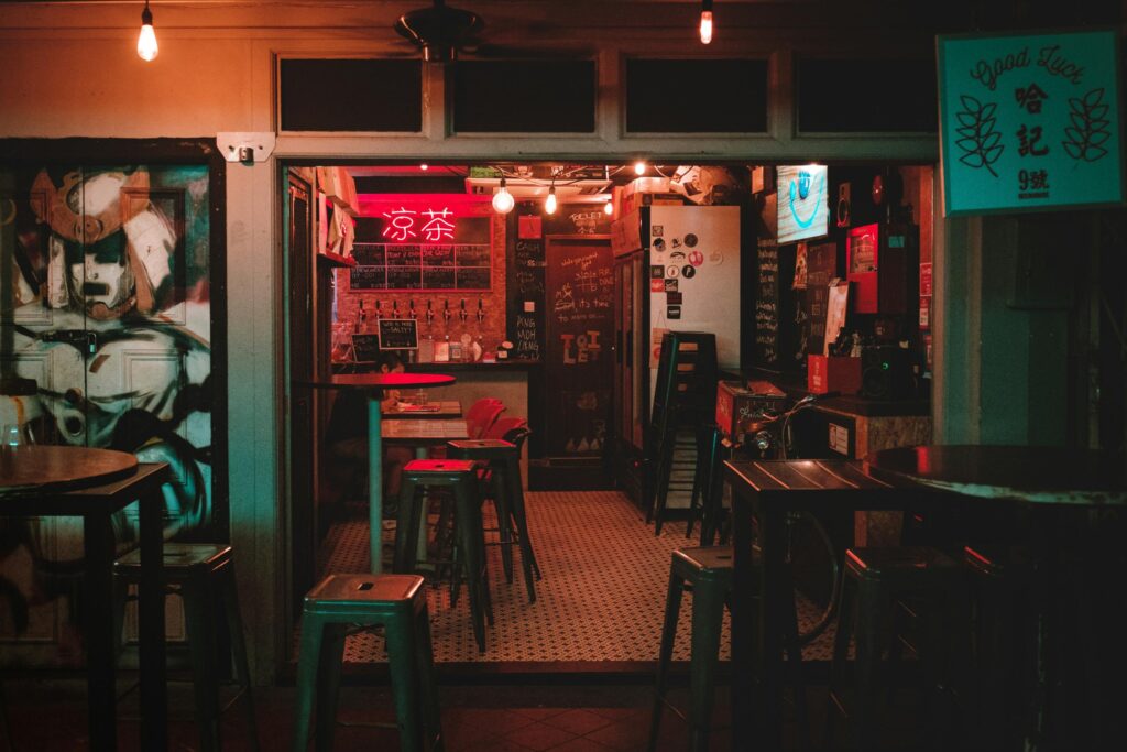 This dimly lit bar features a vibrant mix of eclectic decor, including glowing neon signs, chalkboards, and a large mural on the left. High metal stools surround dark tables, creating a cozy and urban atmosphere reminiscent of a hidden city tavern.
