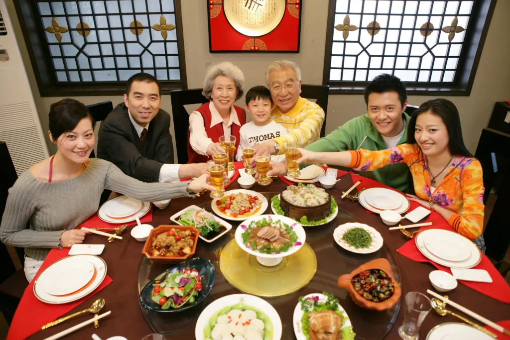 A multi-generational family of seven smiles warmly as they gather around a large round table filled with various Chinese dishes. They are captured in a celebratory moment, raising their glasses together for a toast in a traditional dining setting.