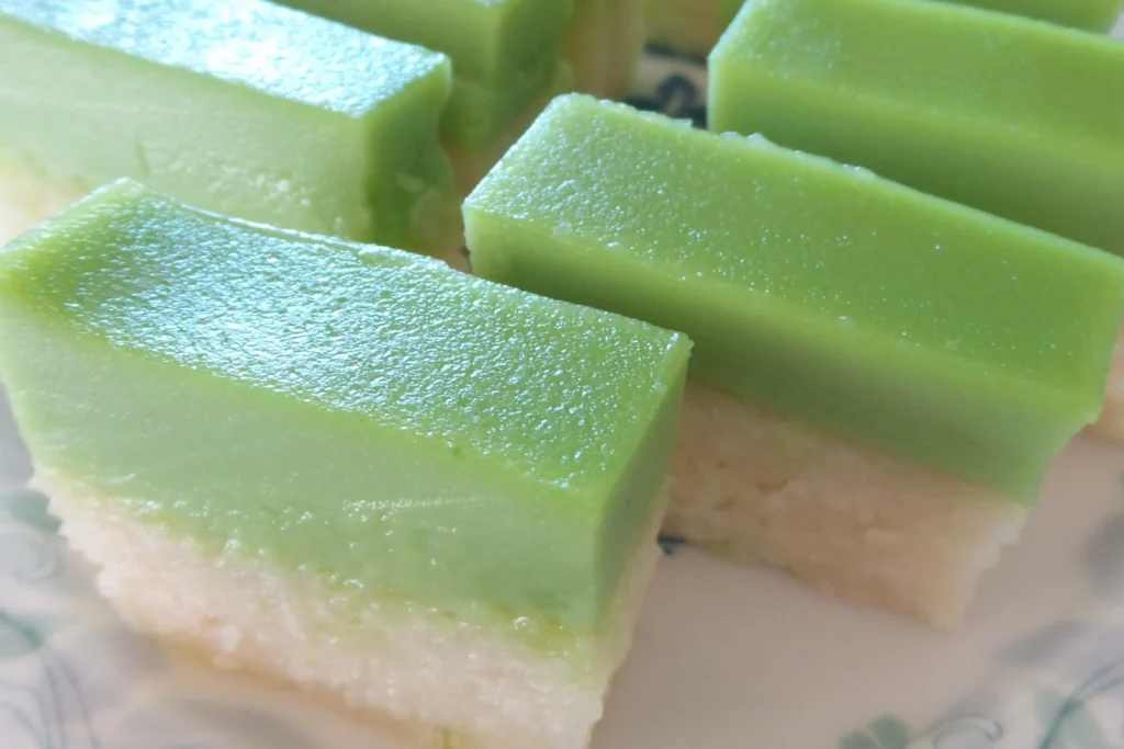 This image features several rectangular slices of Kueh Salat, a traditional Southeast Asian dessert consisting of a vibrant green pandan custard layer atop a white glutinous rice base. The slices are neatly arranged on a patterned plate, showcasing the smooth texture of the custard and the grainy consistency of the steamed rice.