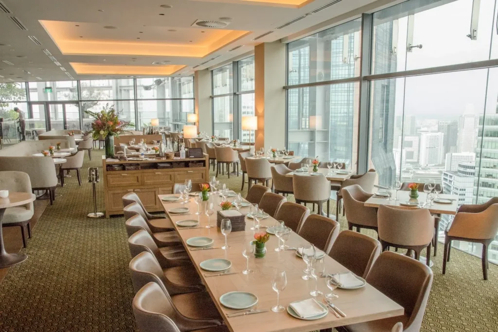 This upscale restaurant dining room features elegant light wood tables and neutral-toned seating arranged against floor-to-ceiling windows with a city skyline view. The space is illuminated by warm recessed ceiling lights and floor lamps, creating a sophisticated and inviting atmosphere.