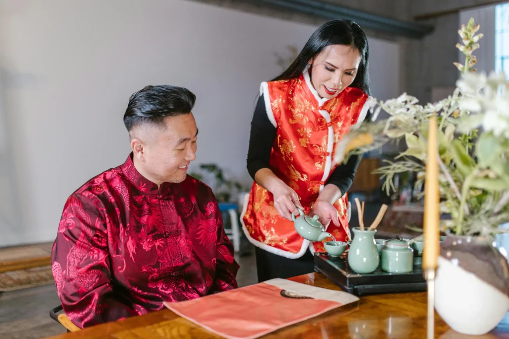 A woman in a red and gold vest is pouring tea from a pale green teapot for a smiling man seated at a wooden table. Both are dressed in traditional Chinese-style attire, creating a warm and cultural atmosphere during their tea service.