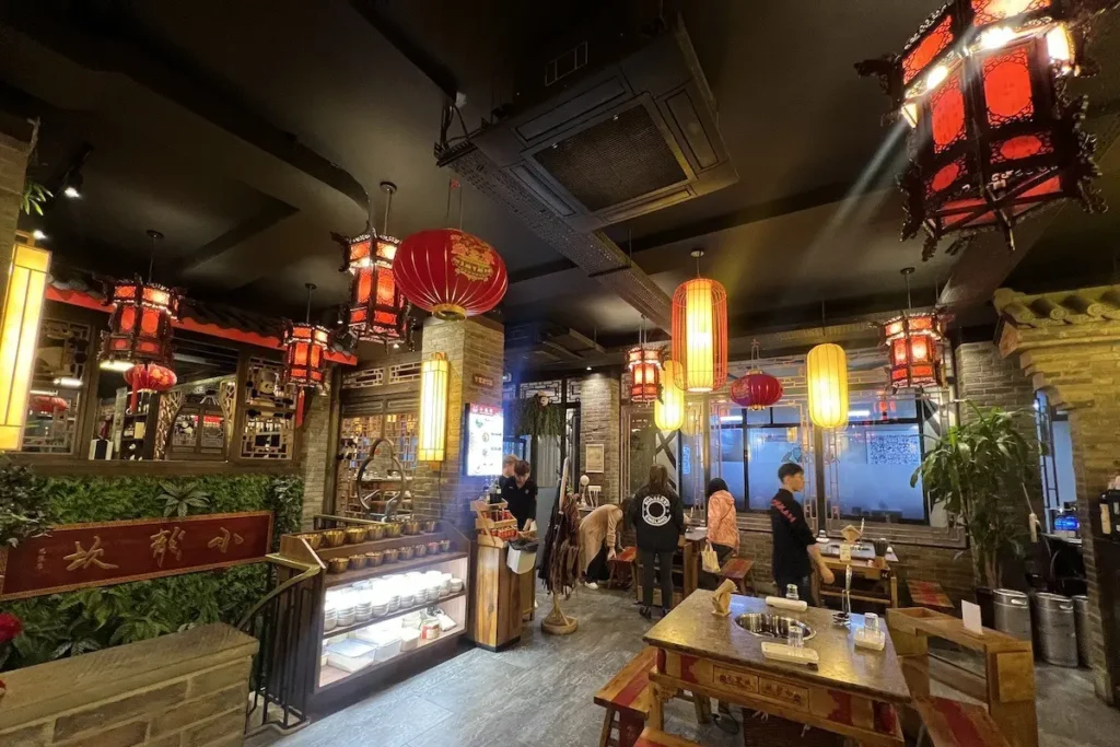 This lively restaurant interior is filled with traditional Chinese lanterns and warm, glowing lights that hang above patrons seated at wooden tables. Brick accents, lush greenery, and ornate woodwork create a cozy, culturally-rich atmosphere for dining.
