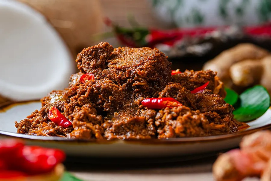 This image features a rich, dark beef rendang garnished with bright red chili slices and served on a simple plate. The background is artfully blurred, highlighting the dish alongside traditional ingredients like fresh coconut and ginger.