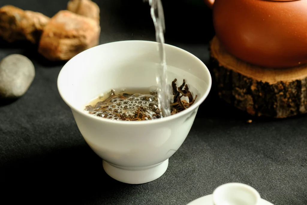 Hot water is poured from above into a white porcelain teacup, causing bubbles to form as it steeps the loose tea leaves. The serene scene is set against a dark background, featuring a clay teapot and rustic wooden accents.