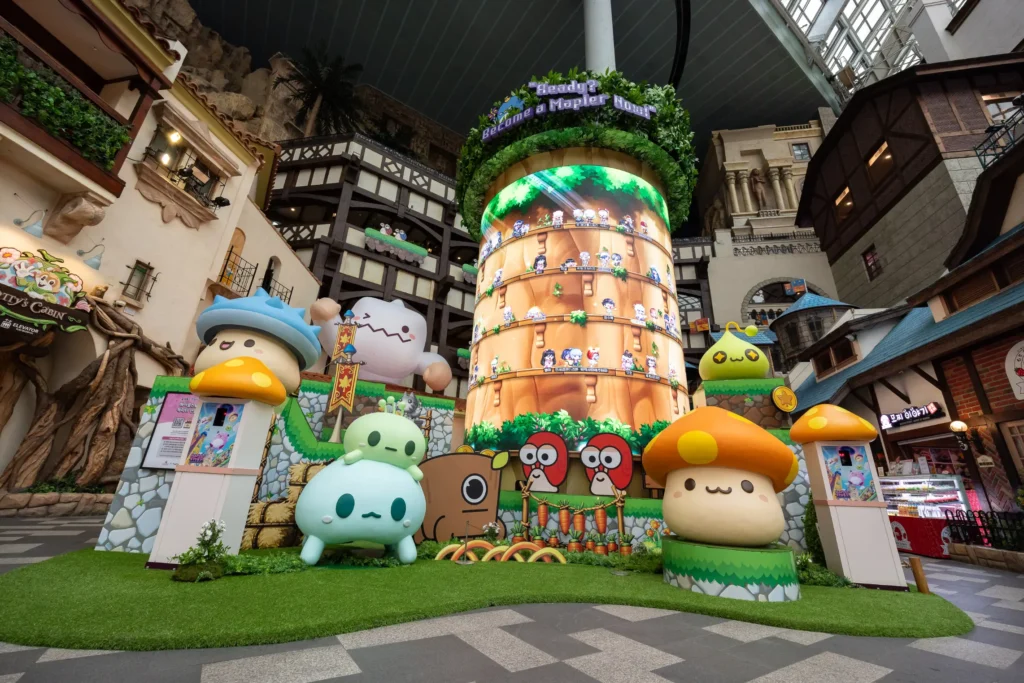 This vibrant indoor display features a large, cylindrical LED screen showcasing characters and settings from the video game MapleStory. Surrounding the screen are various colorful, oversized statues of the game's iconic monsters, including Mushrooms and Slimes, set within a whimsical themed environment.
