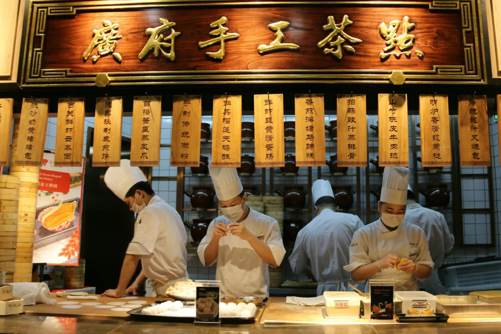 Several chefs in white uniforms and face masks are meticulously preparing dim sum behind a wooden counter. Above them hangs a large, ornate sign and several wooden menu plaques featuring gold Chinese calligraphy.