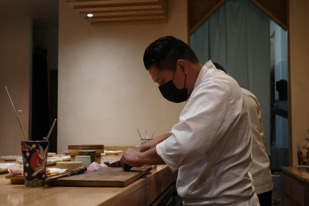 A chef in a white uniform and black face mask is captured in profile, focused on slicing ingredients on a wooden cutting board. The warm, minimalist interior of the sushi bar features clean lines and traditional accents, highlighting the quiet intensity of the preparation.