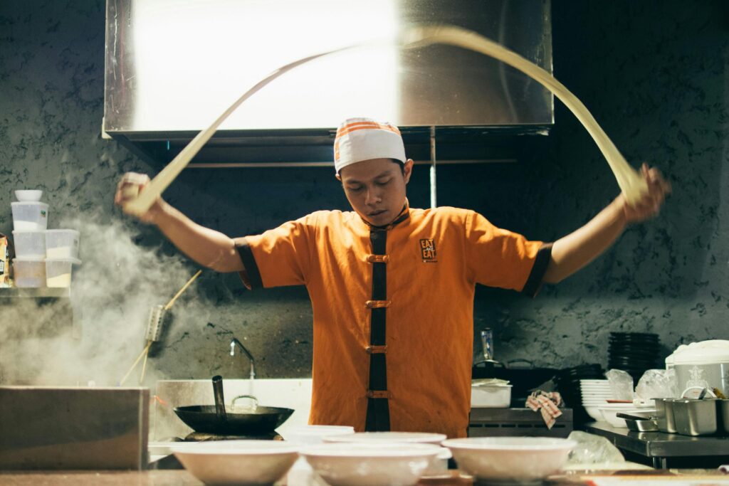 A chef in an orange uniform and white cap skillfully stretches a long strand of noodle dough into a wide arc over a kitchen counter. The bustling kitchen scene is filled with steam, cooking equipment, and white bowls prepared for serving.
