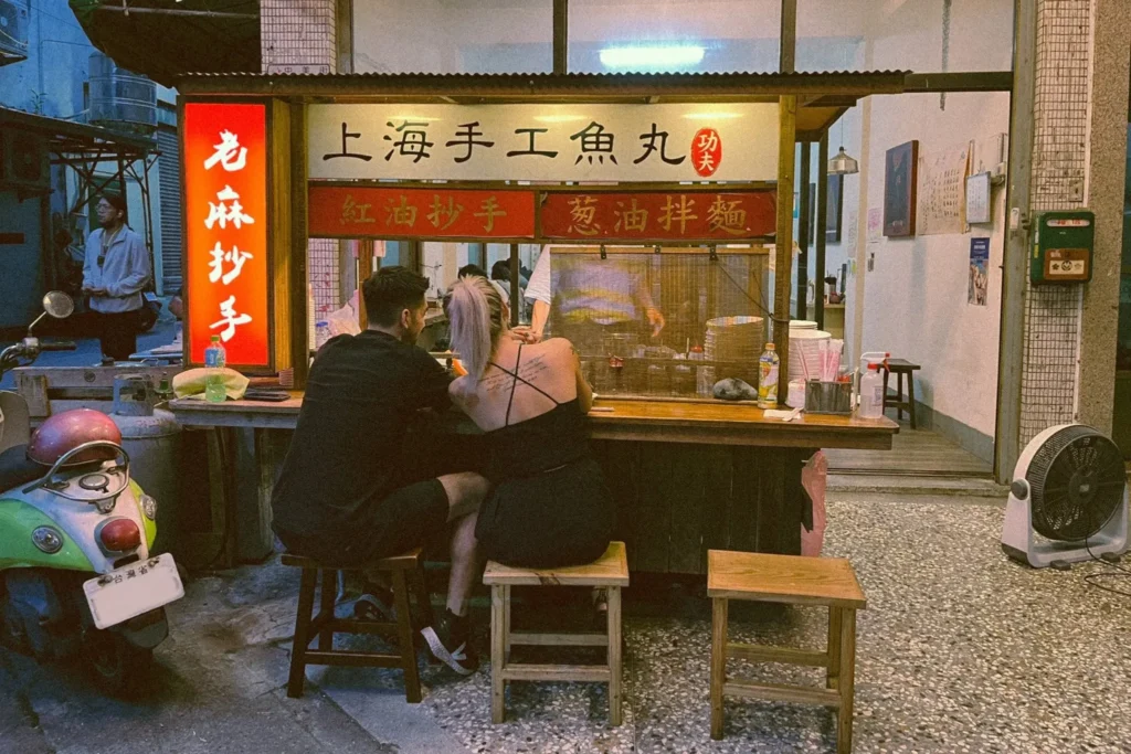 A couple sits on wooden stools at a small outdoor food stall, enjoying a meal under the warm glow of traditional signage. The shop features handwritten menus for Shanghai-style fish balls and spicy wontons, creating an intimate street-side dining atmosphere.