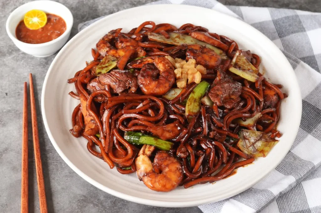 The image features a plate of Hokkien Mee, consisting of thick, dark-sauced noodles tossed with prawns, pork, cabbage, and crispy lard. It is served with a side of sambal chili topped with a calamansi lime, set against a grey background with a checkered cloth and wooden chopsticks.