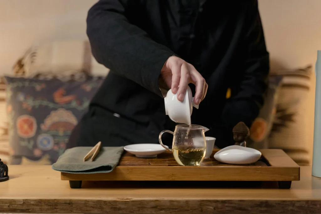 A person in dark clothing is shown pouring tea from a white ceramic vessel into a small glass pitcher on a wooden tea tray. The serene scene features various traditional tea accessories, including a tea pet, tongs, and small dishes arranged for a formal tea ceremony.