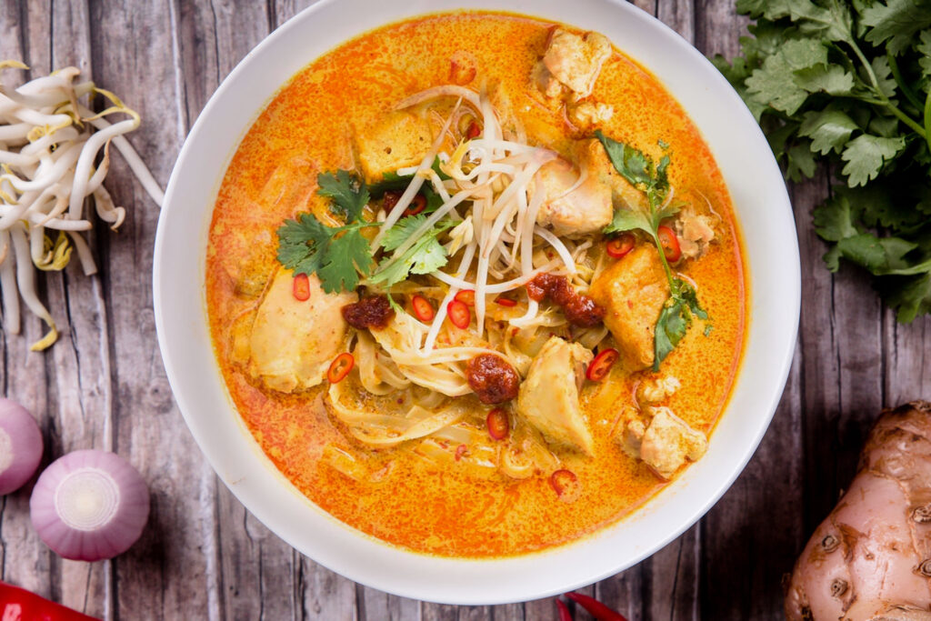 This overhead shot features a vibrant bowl of laksa filled with noodles, tofu puffs, and chicken in a rich, orange coconut curry broth. The dish is beautifully garnished with bean sprouts, fresh cilantro, and sliced red chilies, with raw ingredients like galangal and shallots scattered on the wooden surface nearby.