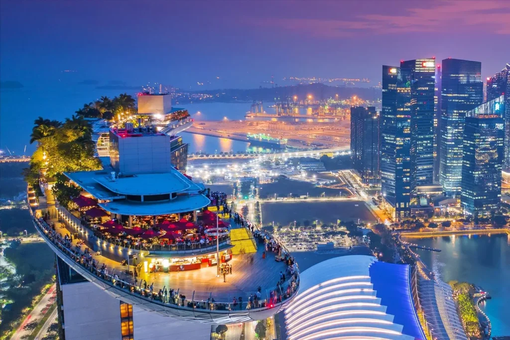 An aerial evening shot captures the illuminated Sands SkyPark observation deck at Marina Bay Sands, overlooking the vibrant city lights and harbor of Singapore. Crowds of people gather on the cantilevered deck to enjoy panoramic views of the surrounding skyscrapers and the glowing waterfront.